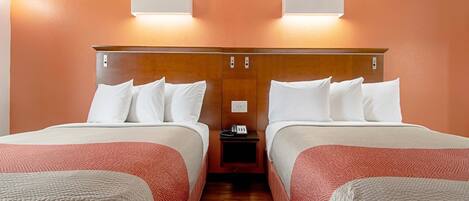A hotel room with two beds, each with white bedding and a red patterned blanket. There are wall-mounted light fixtures above the beds, and a small table with a phone between them.