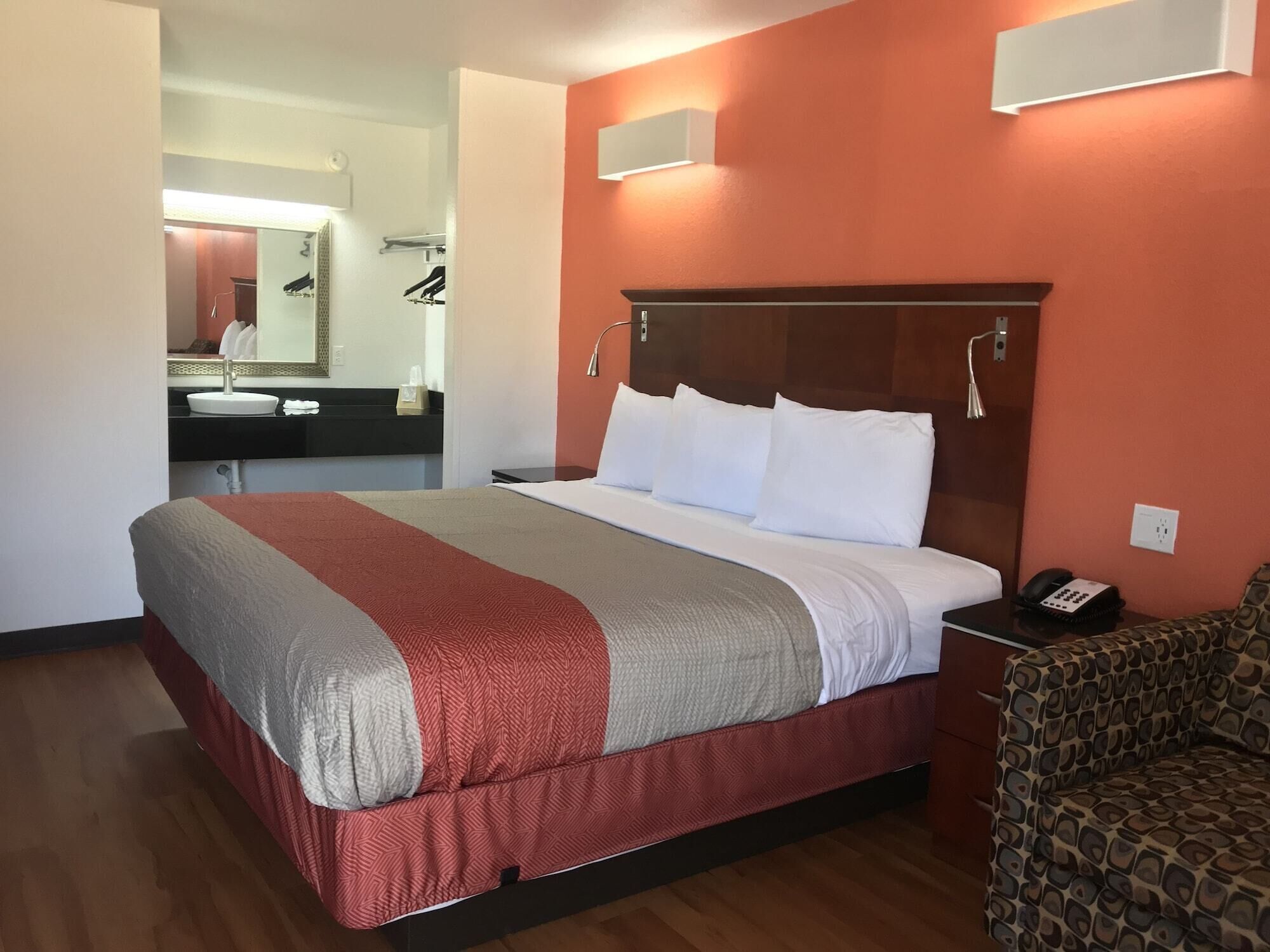 Una habitación de hotel con una cama, una mesita de noche con teléfono, una silla, un baño con lavamanos y espejo, y un aplique de pared.
