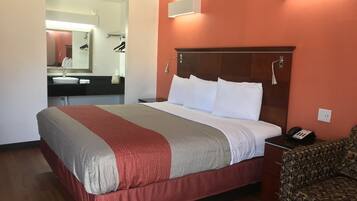 Una habitación de hotel con una cama, una mesita de noche con teléfono, una silla, un baño con lavamanos y espejo, y un aplique de pared.