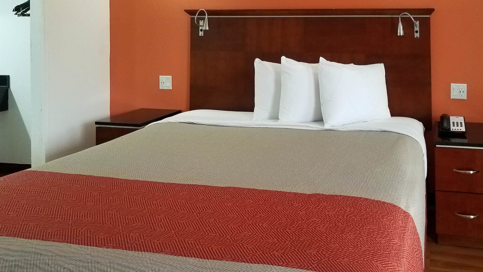 Ein Hotelzimmer mit einem großen Bett, einem hölzernen Kopfteil, weißen Kissen, einer rot-beigen Bettdecke, einem Nachttisch mit Telefon und einer Holzkommode.