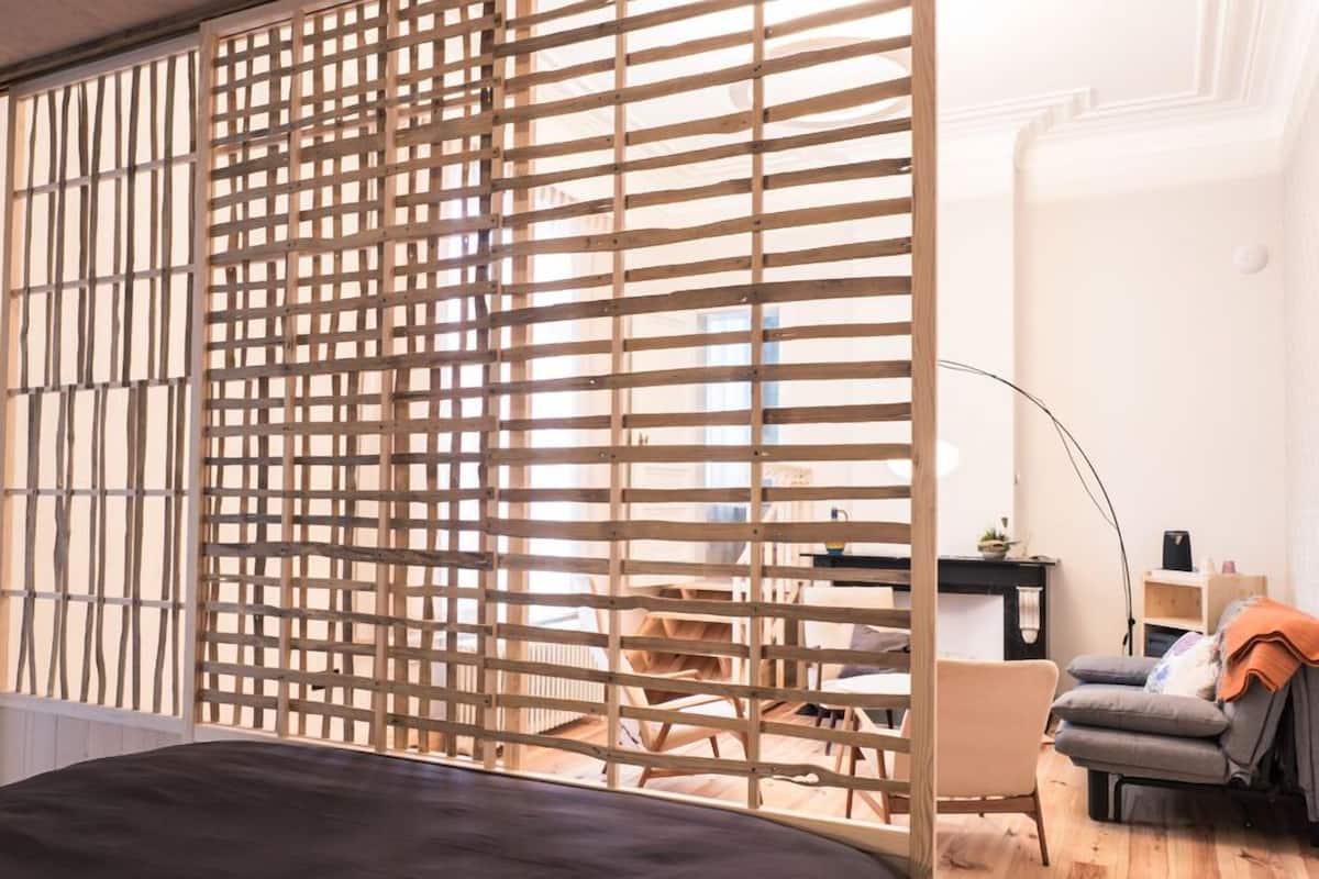 A room with a bed, a desk, and a chair. There is a window with a wooden slat screen.