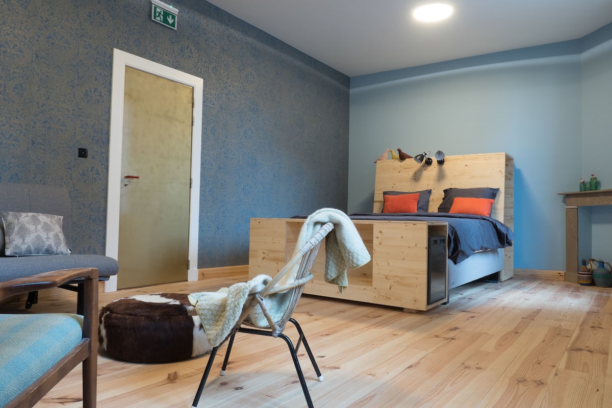 A room with a wooden bed, a chair with a blanket, a sofa, and a wooden floor.