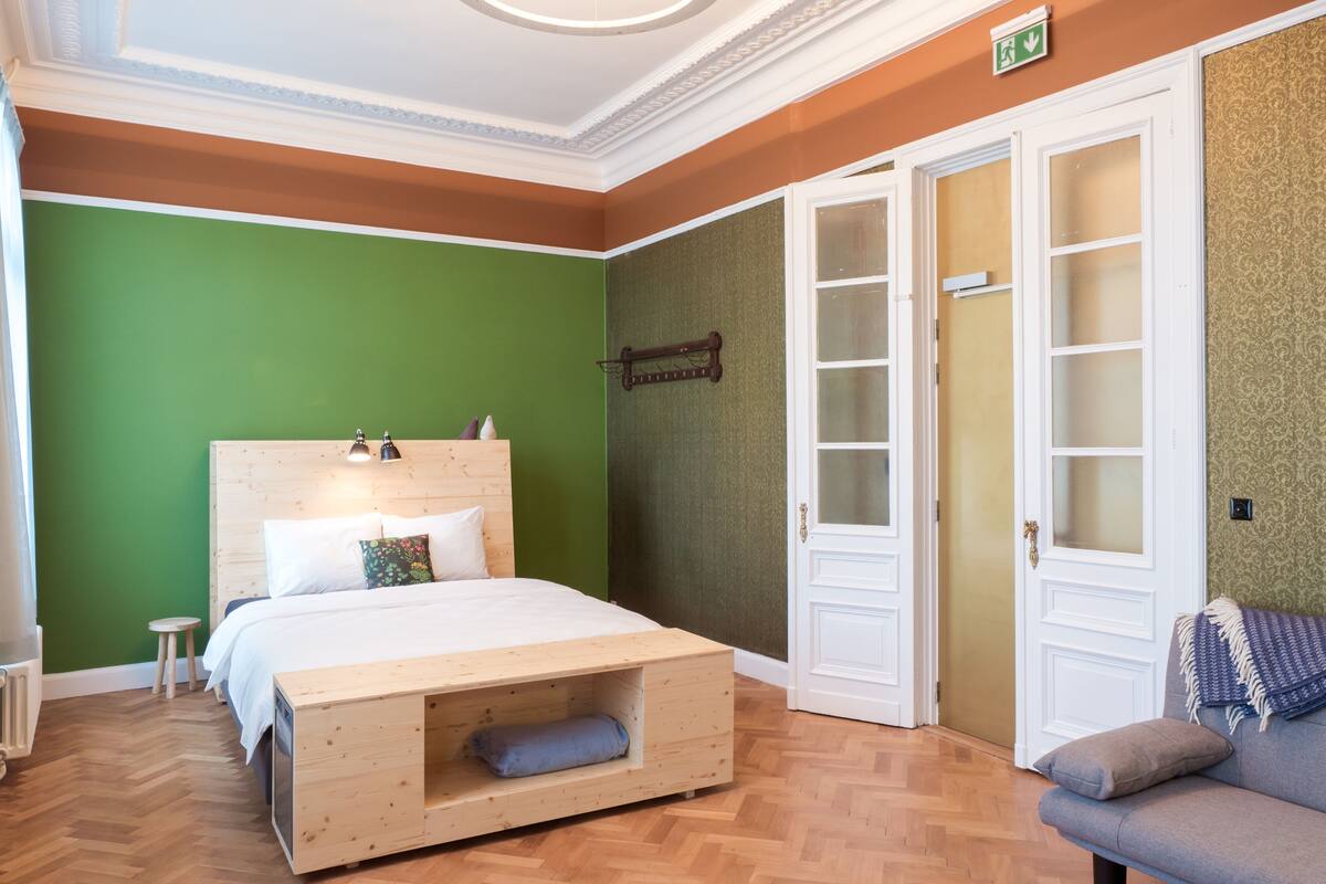 A room with a wooden bed, green walls, a herringbone floor, a white door, and a small stool.