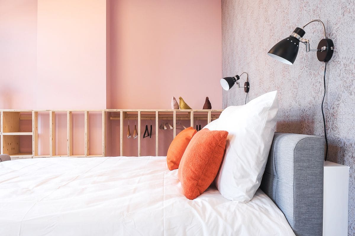 A neatly made bed with white and orange pillows, a grey headboard, and a wall-mounted lamp.