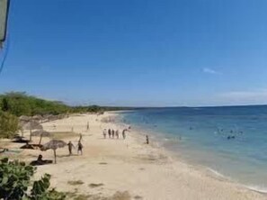A beach with clear blue water, people walking along the shore, and some vegetation on the left. - Hostal plaza (Cienfuegos)