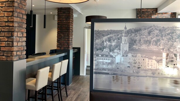 A modern interior with a bar area, a large window with a cityscape view, and exposed brick walls. - Rafaela Hotel Heidelberg (Heidelberg)