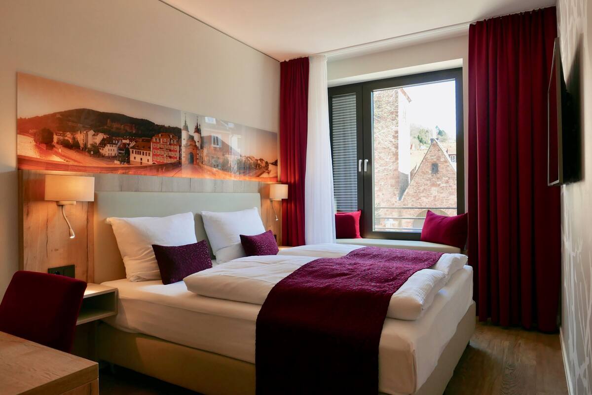 A hotel room with a large bed, a painting on the wall, a window with red curtains, and a TV on the wall.