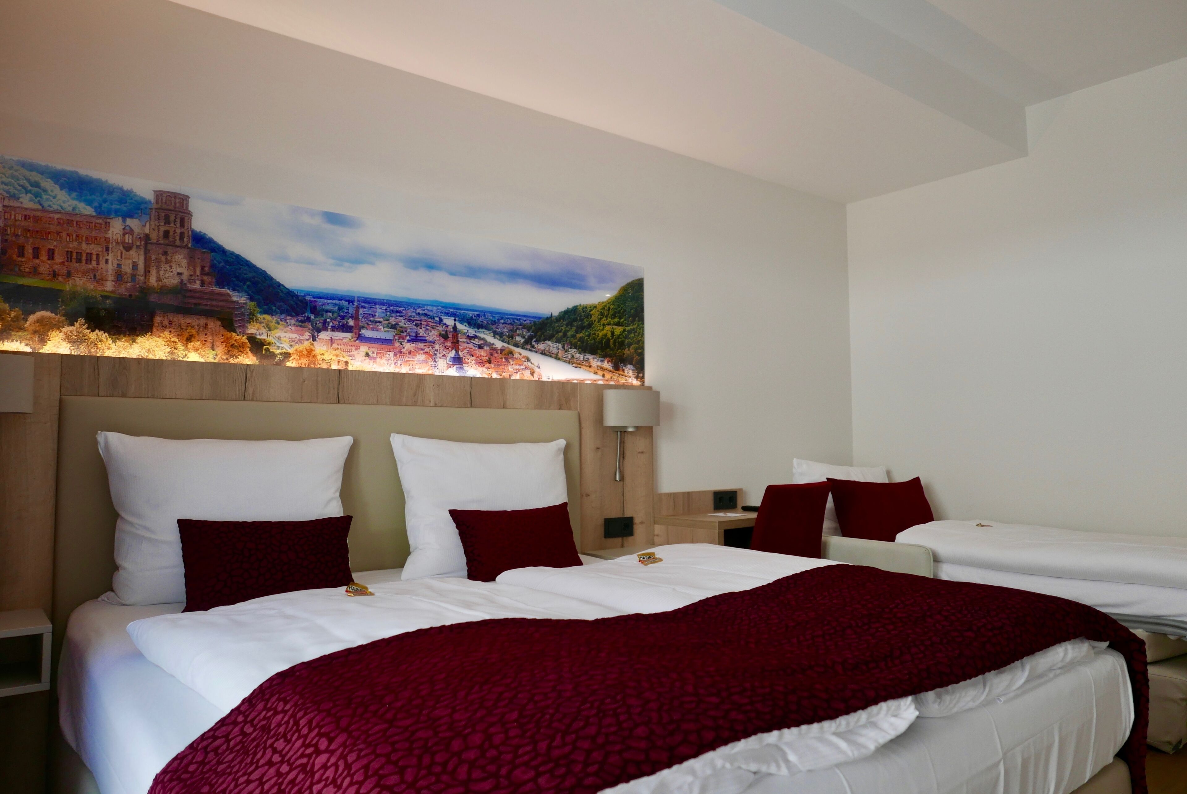A hotel room with a large bed, a headboard, and a wall-mounted painting of a cityscape.