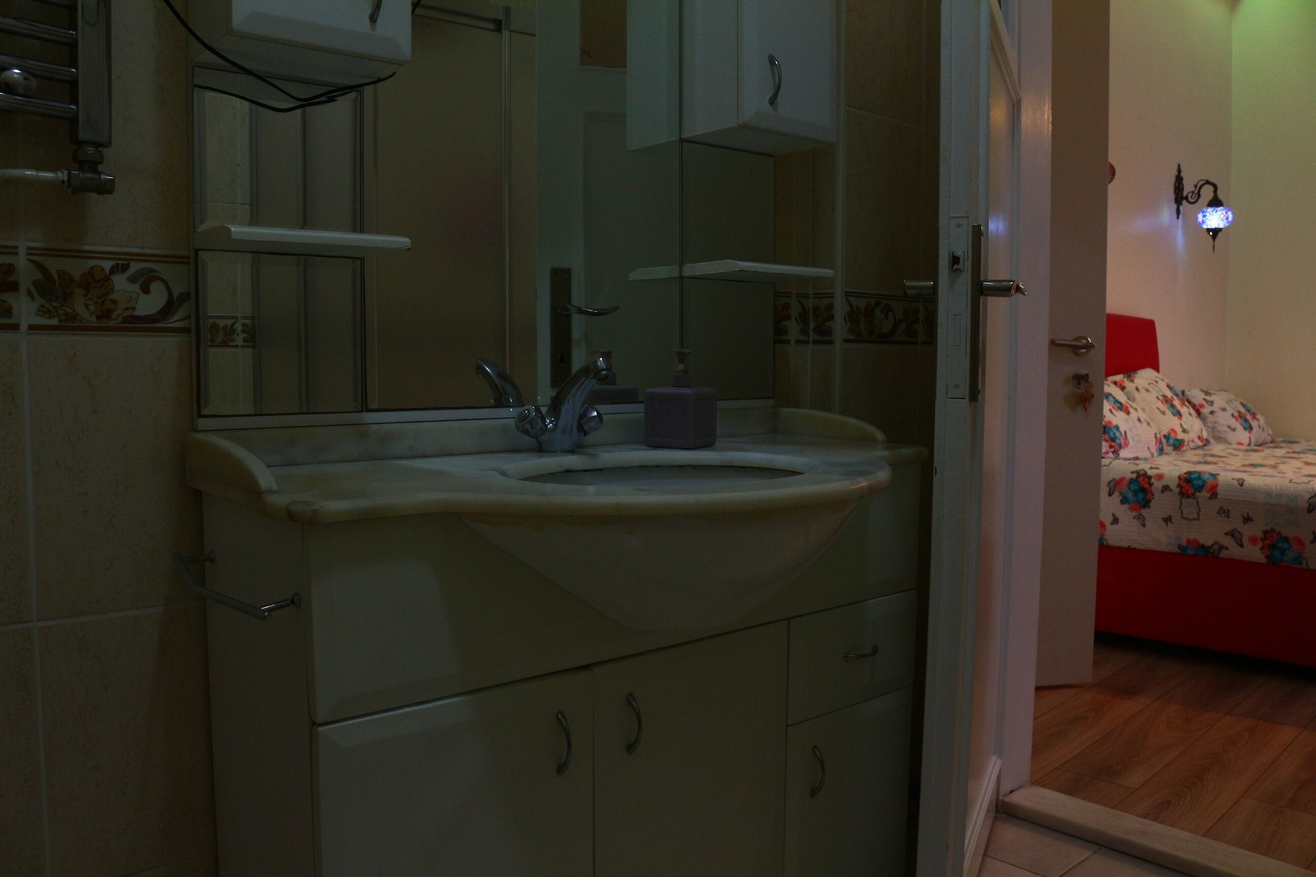 A bathroom with a sink, mirror, and cabinets. There is a door leading to a bedroom with a bed.