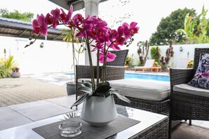 A patio area with a white vase containing pink orchids, wicker furniture, and a pool in the background.