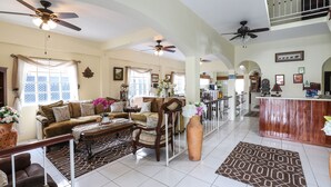 A spacious living room with a high ceiling, multiple ceiling fans, a large wooden dining table, and a kitchen area with a bar counter.