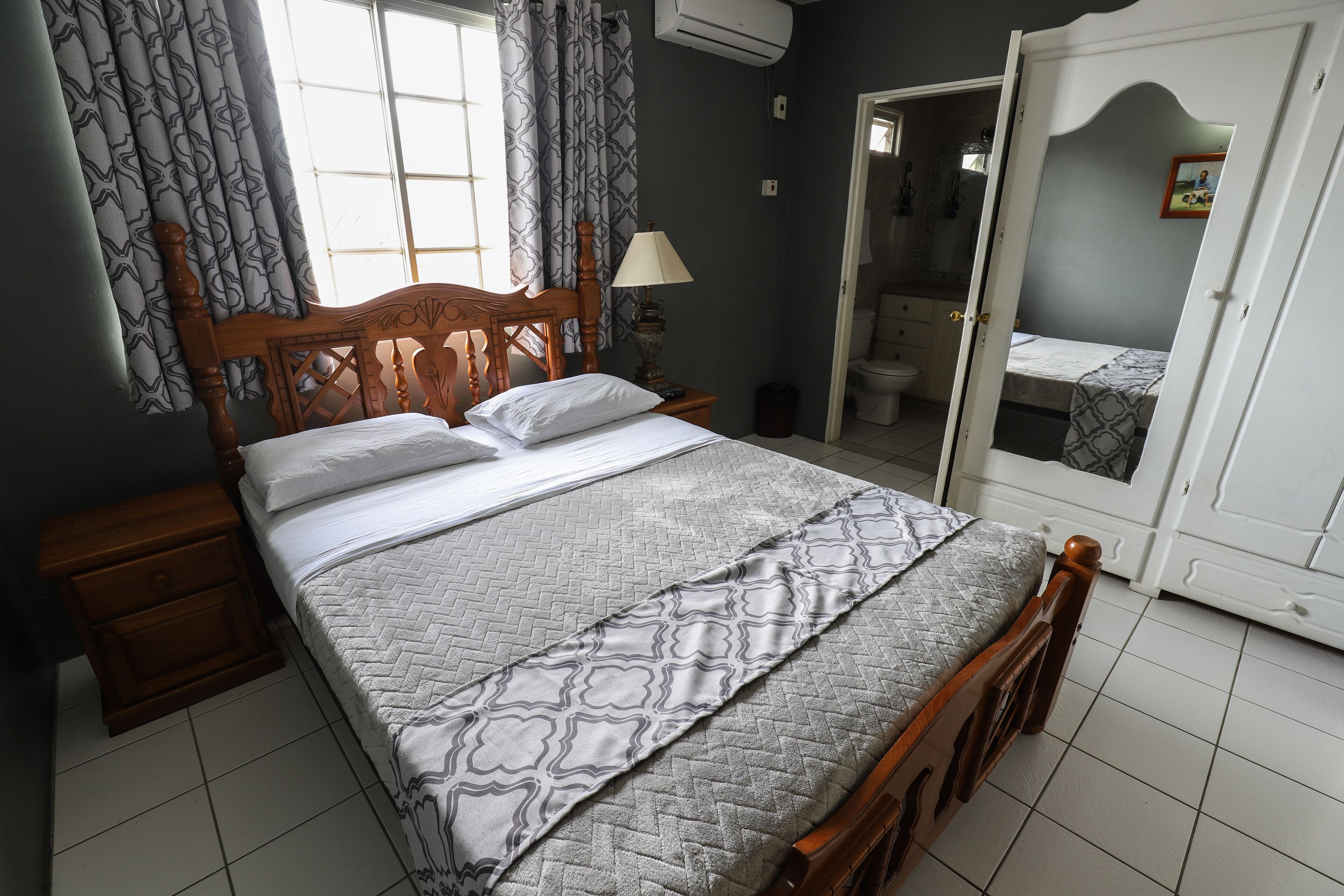 A bedroom with a wooden bed, a bedside table, a nightstand, a lamp, and a bathroom visible through an open door.