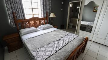 Quarto com cama de madeira, mesa de cabeceira, luminária e banheiro visível por uma porta aberta.