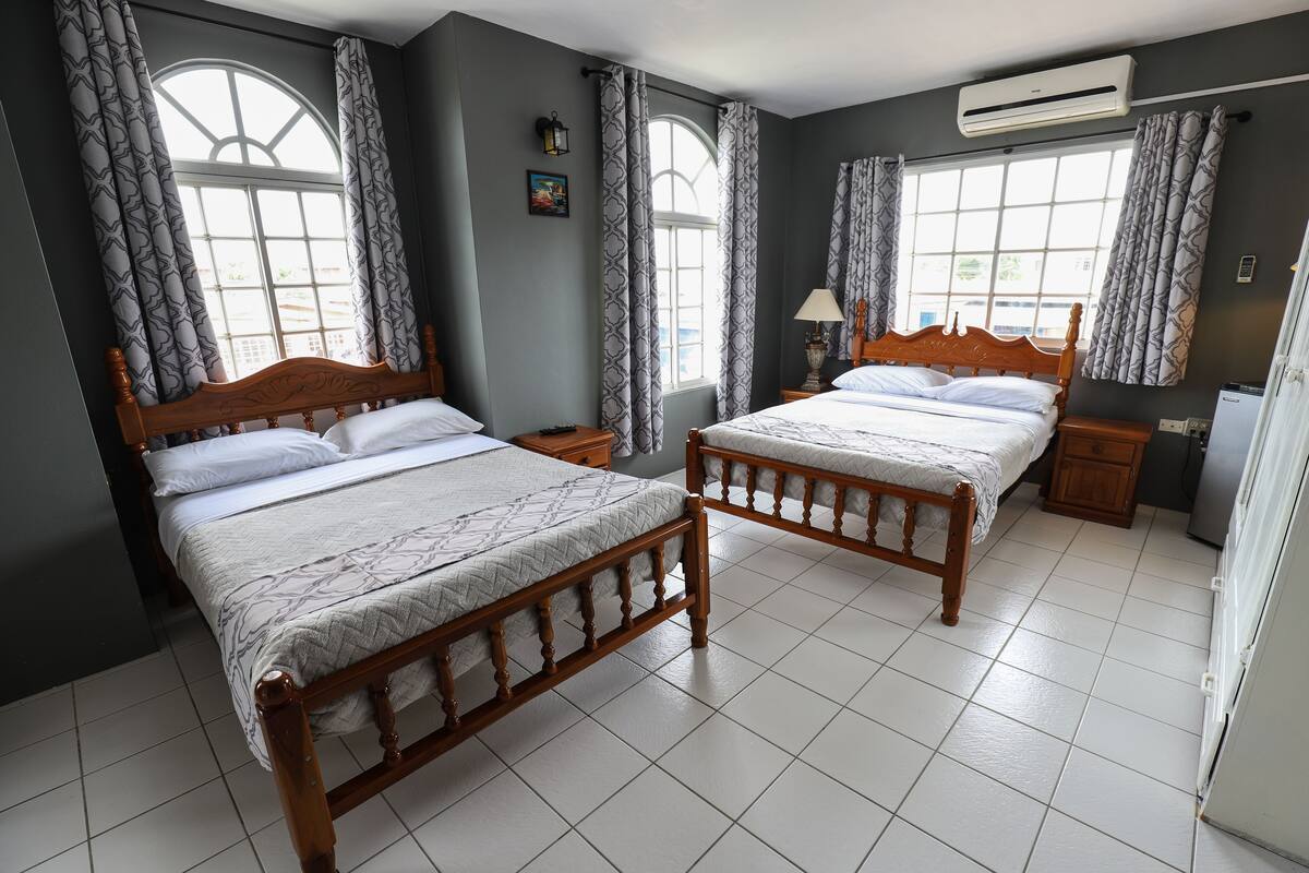 A hotel room with two wooden beds, a large window with curtains, a wall-mounted air conditioner, and a small bedside table with a lamp.