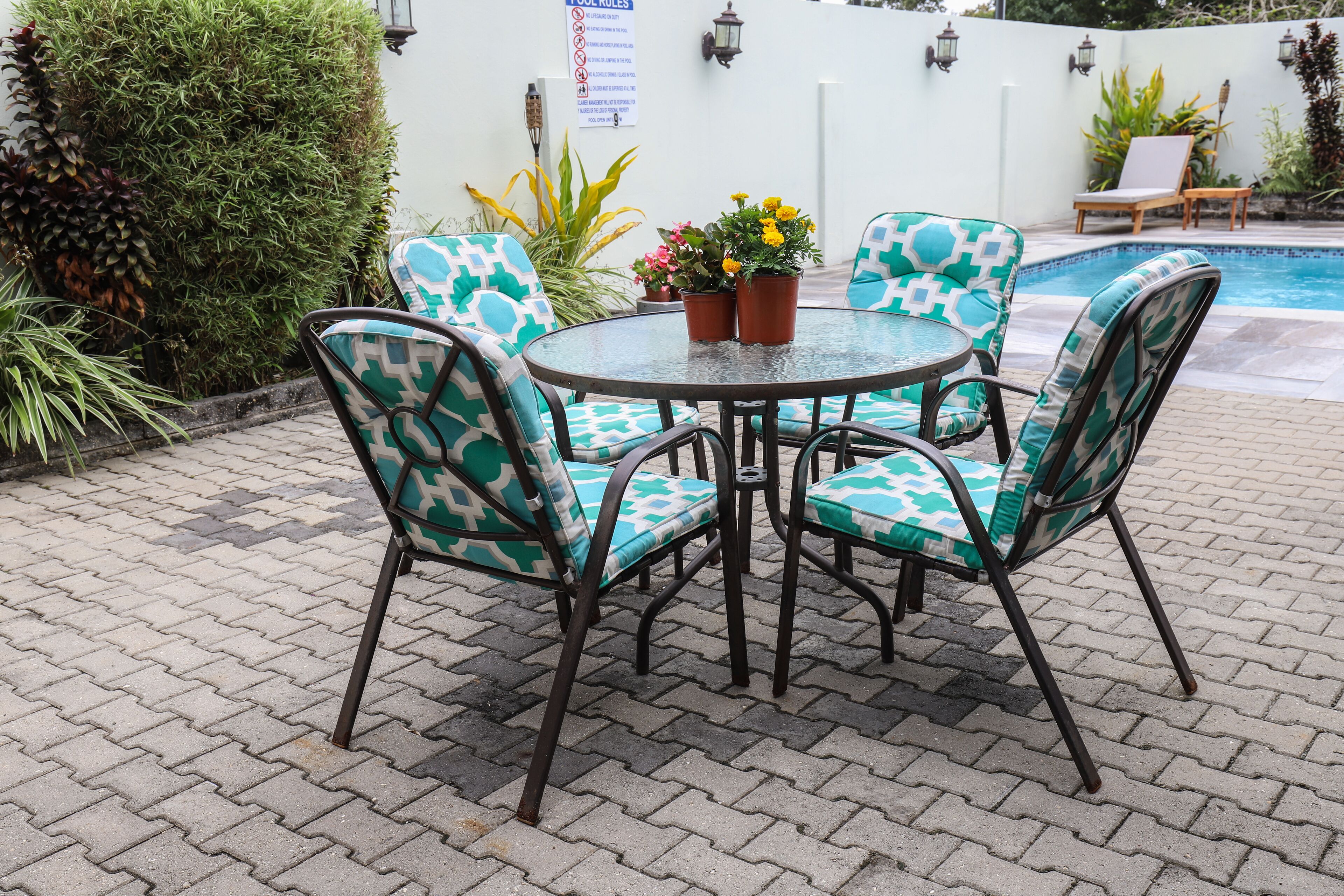 A round table with four chairs, each with a geometric pattern, surrounded by potted plants and a pool in the background.