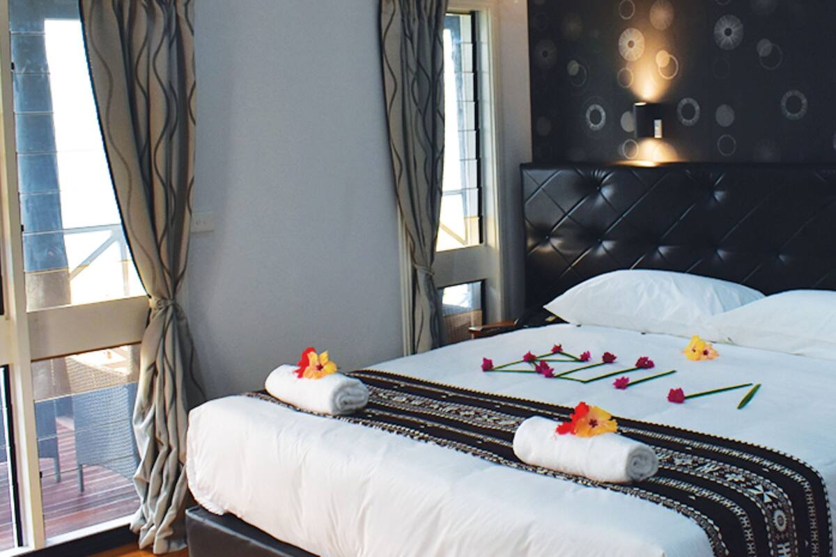 A neatly made bed with white linens and decorative flowers, a dark headboard, and a view of the outdoors through a window with curtains.