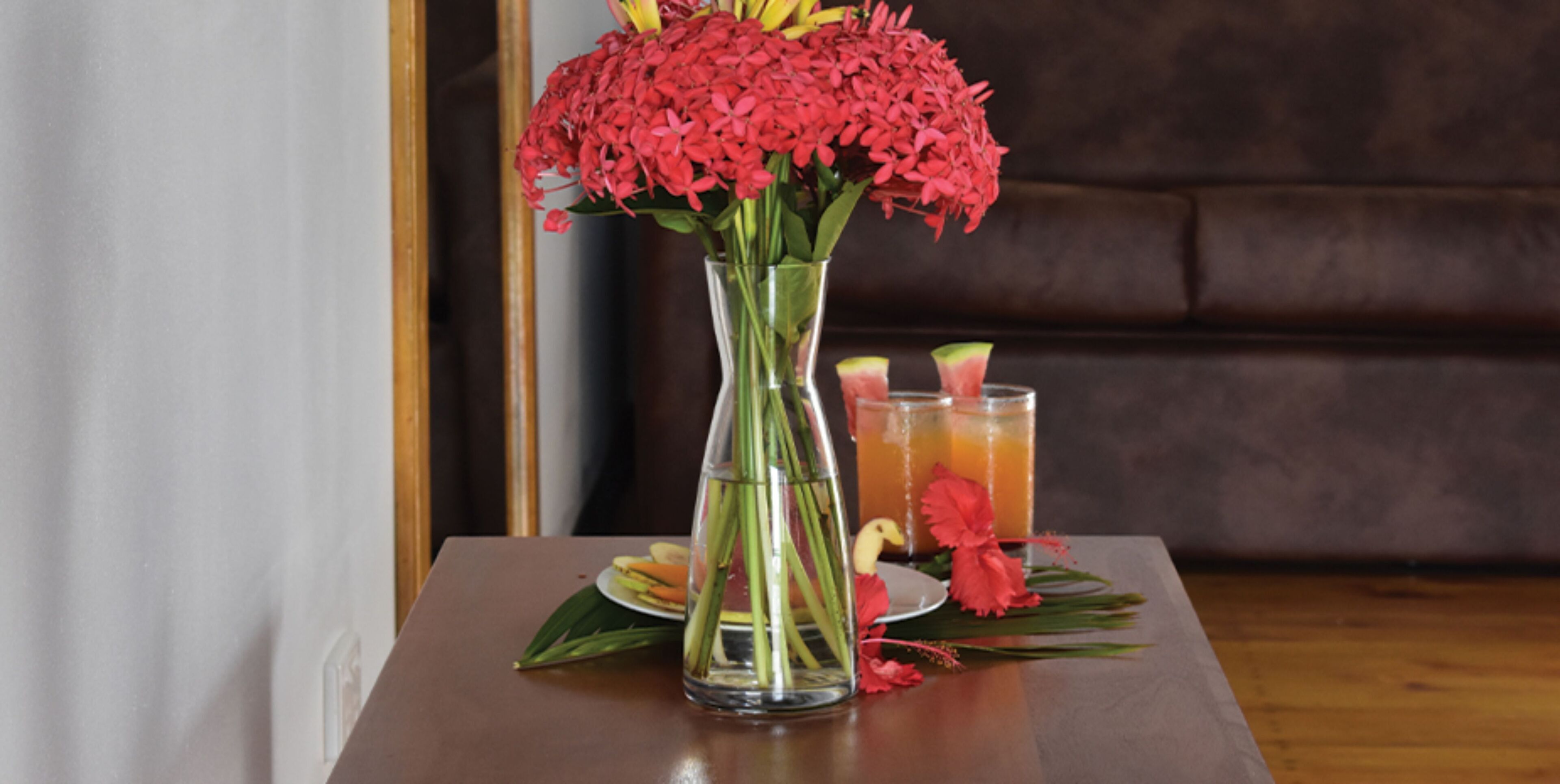 A table with a vase of red flowers, a glass of orange juice, and a plate with food.