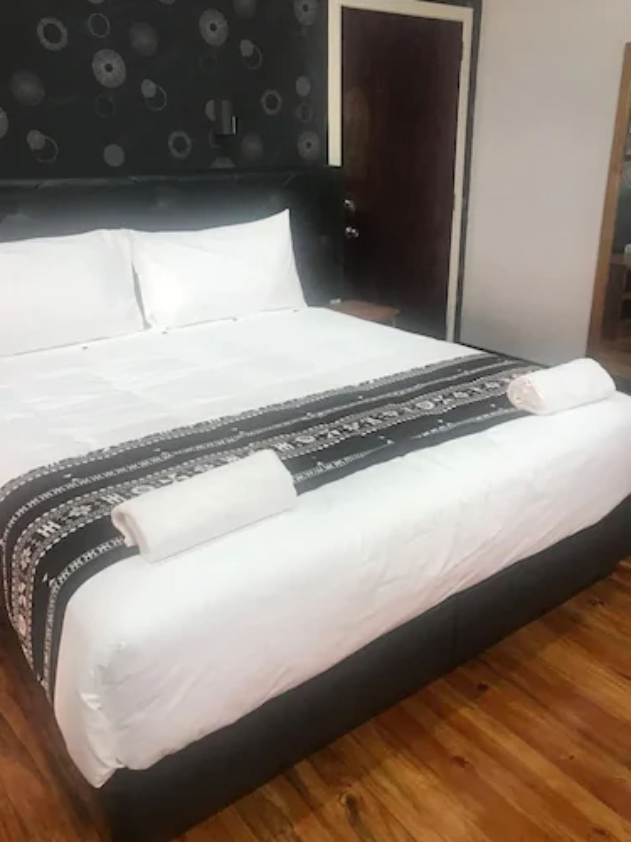 A neatly made bed with white linens and a patterned blanket, a dark headboard with circular designs, and a wooden floor.