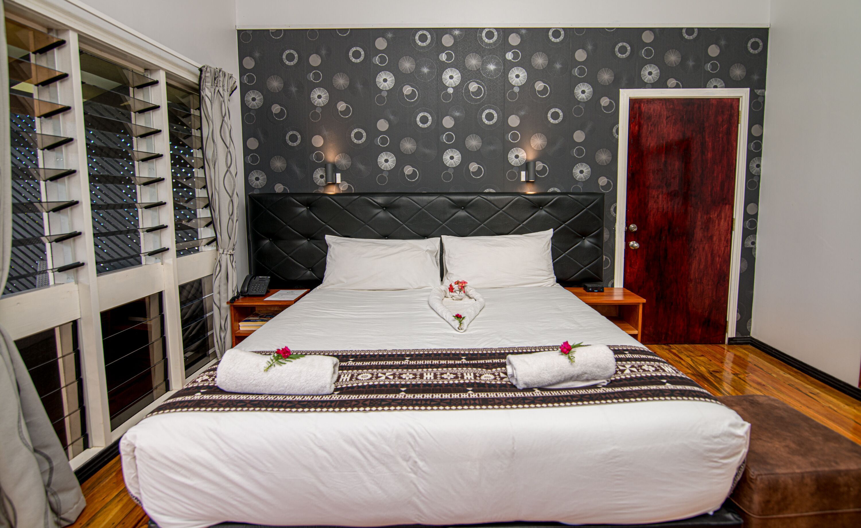 Habitación de hotel con una cama grande, cabecera estampada oscura, mesita de noche de madera, un aparador, un teléfono y una puerta con marco rojo.