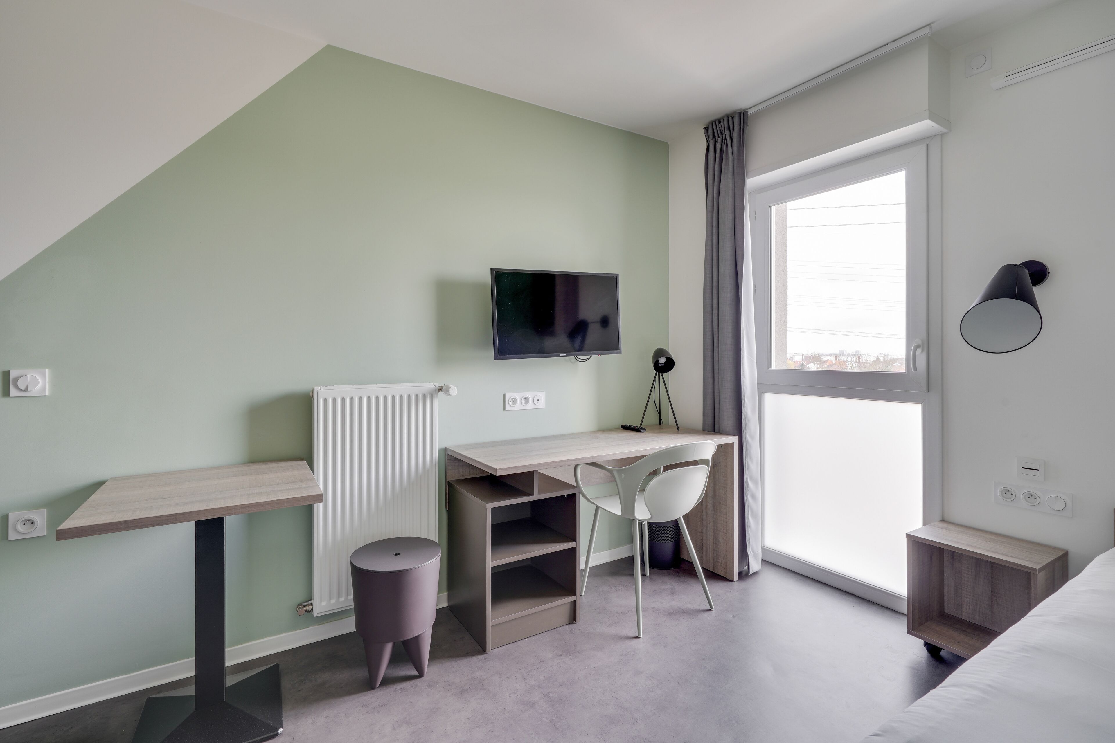 Een moderne slaapkamer met een schuine kap, een bureau met stoel, een televisie, een nachtkastje en een groot raam.