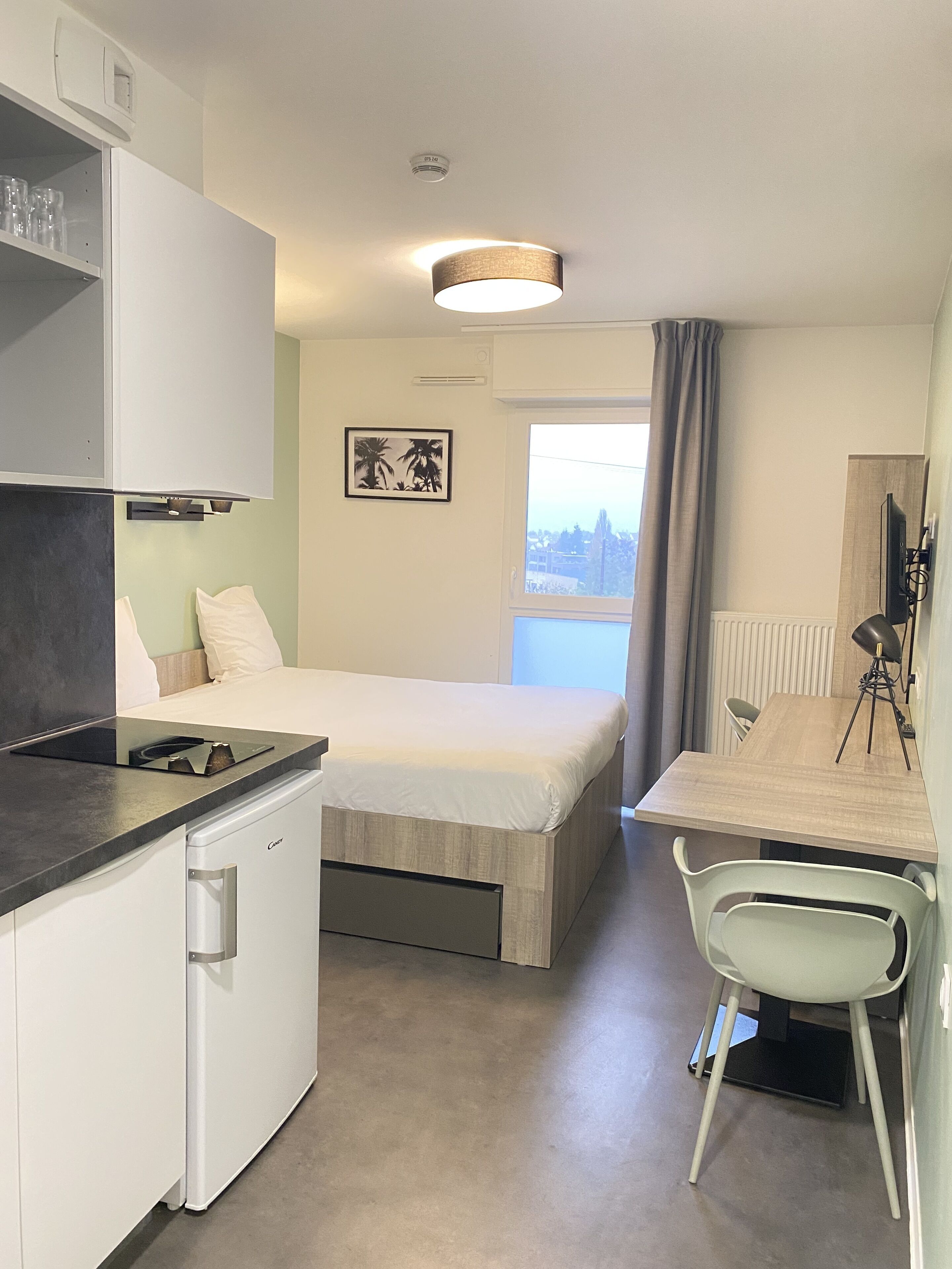 Une chambre d’hôtel compacte comprenant un lit, une petite kitchenette, une table à manger avec une chaise et une fenêtre donnant sur un paysage agréable.