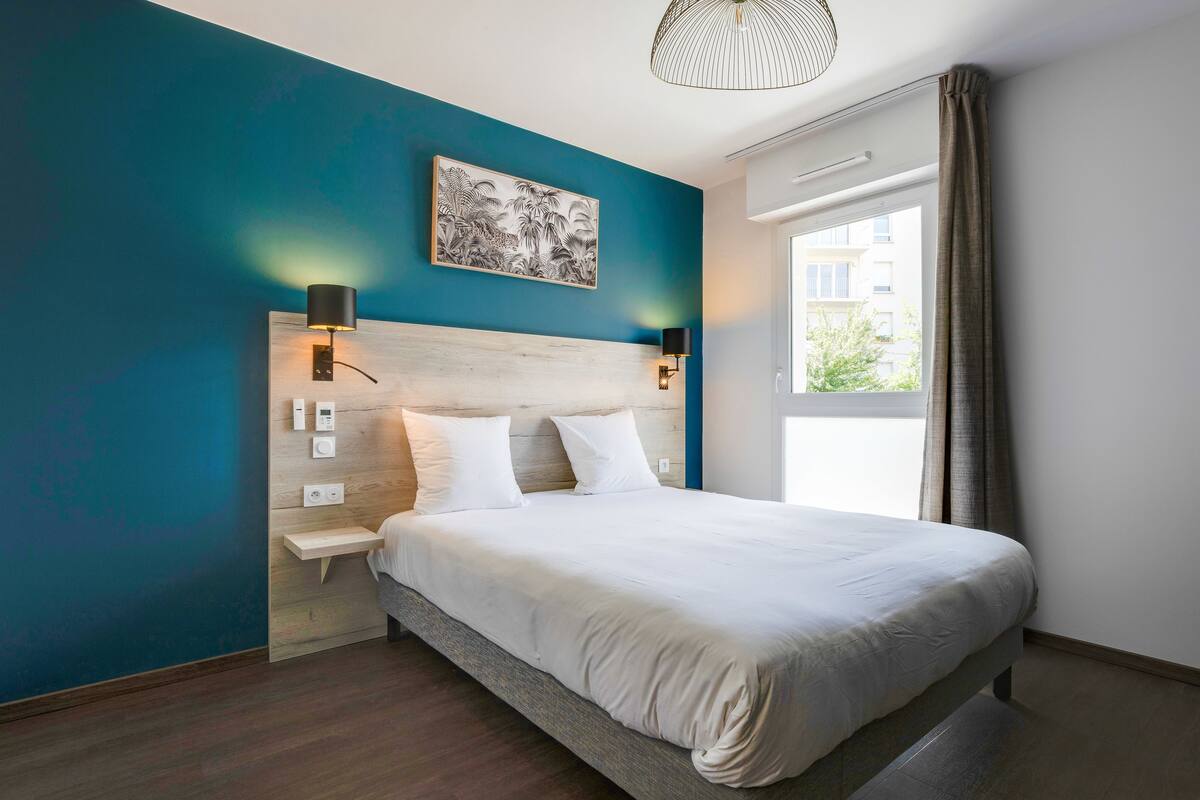 A modern hotel room with a large bed, a wooden headboard, a wall-mounted artwork, a nightstand, and a window with curtains.