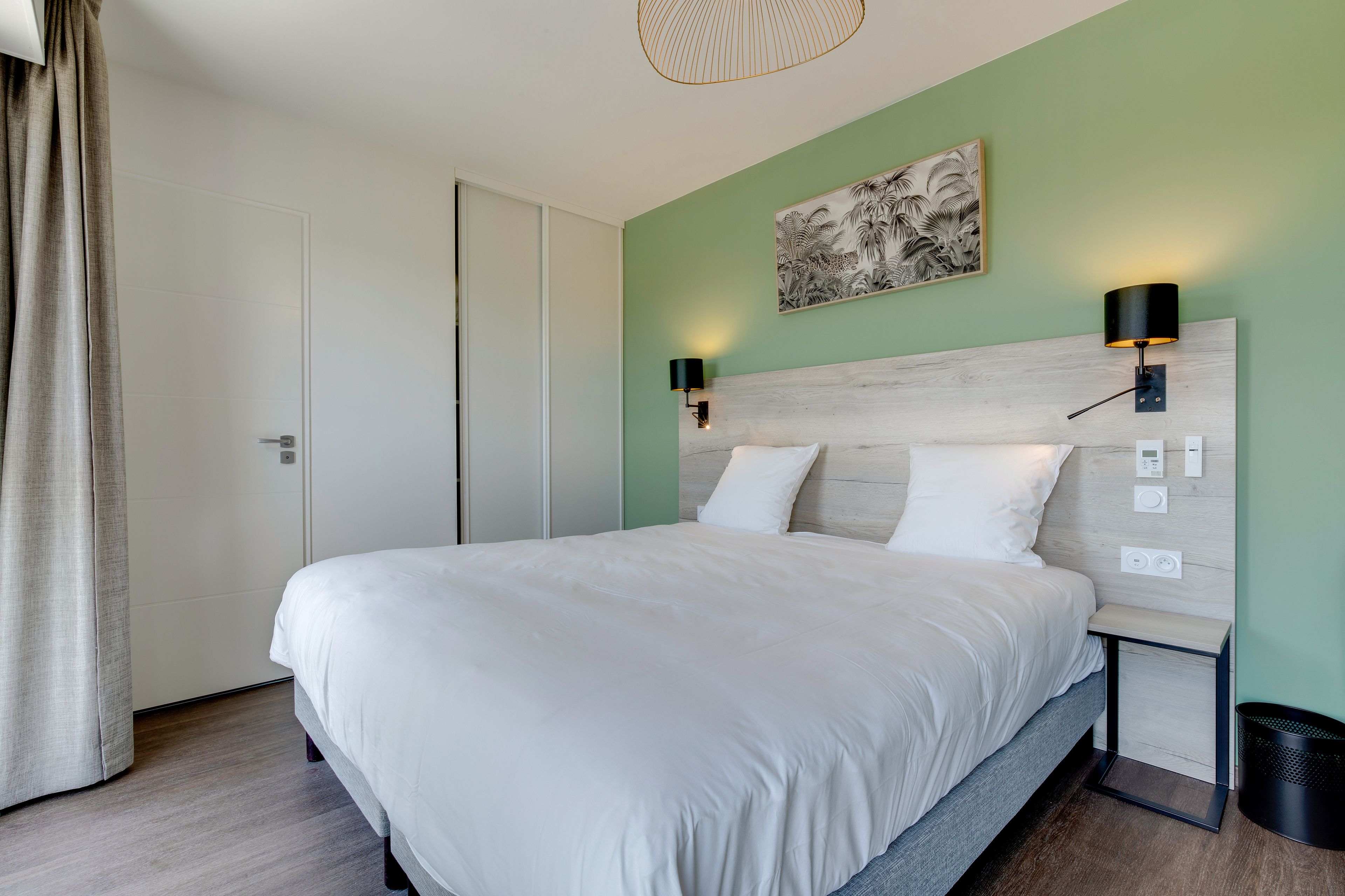 A neatly made bed in a room with a green wall, a wooden headboard, and two wall-mounted lamps.