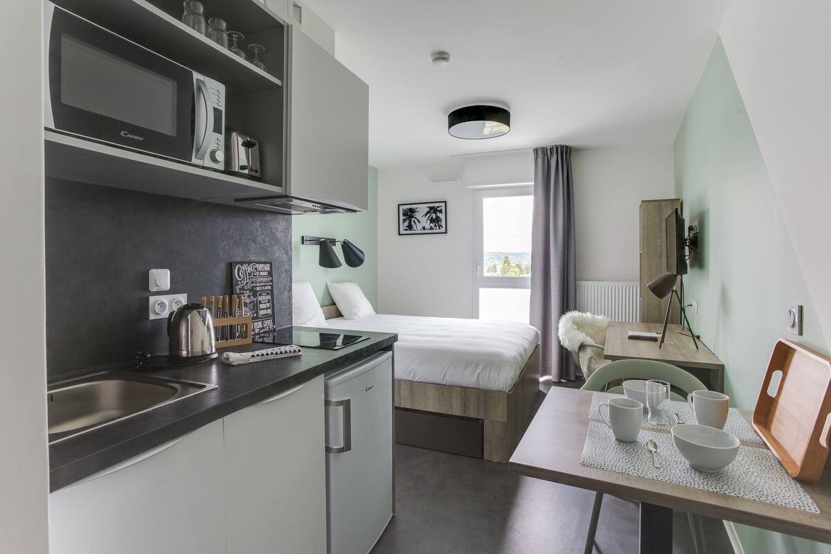 A compact hotel room with a kitchenette, a bed, a dining table with chairs, and a view of the outdoors.