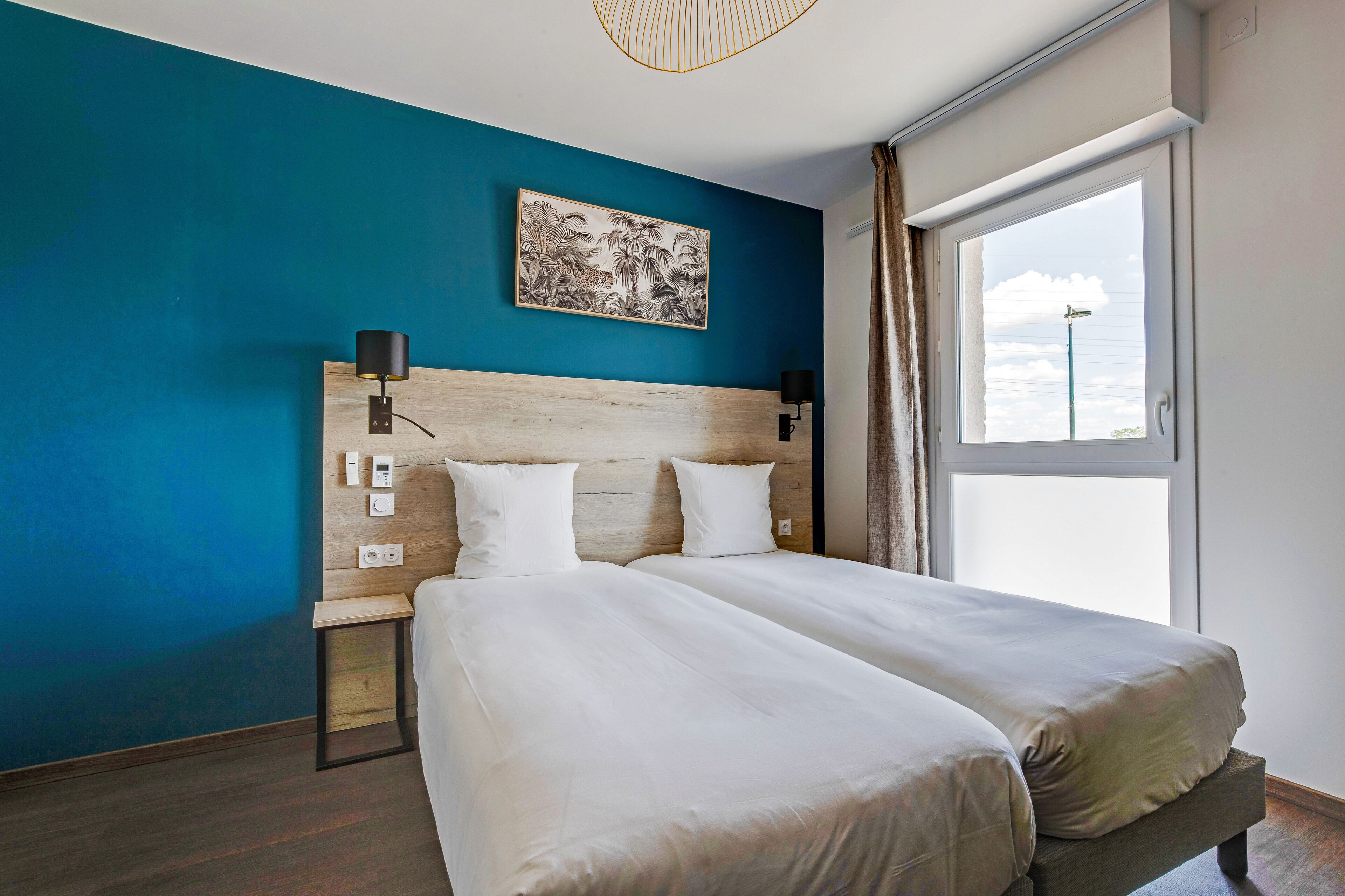 A double bed with a wooden headboard, bedside lamps, a wall-mounted artwork, and a window with curtains.