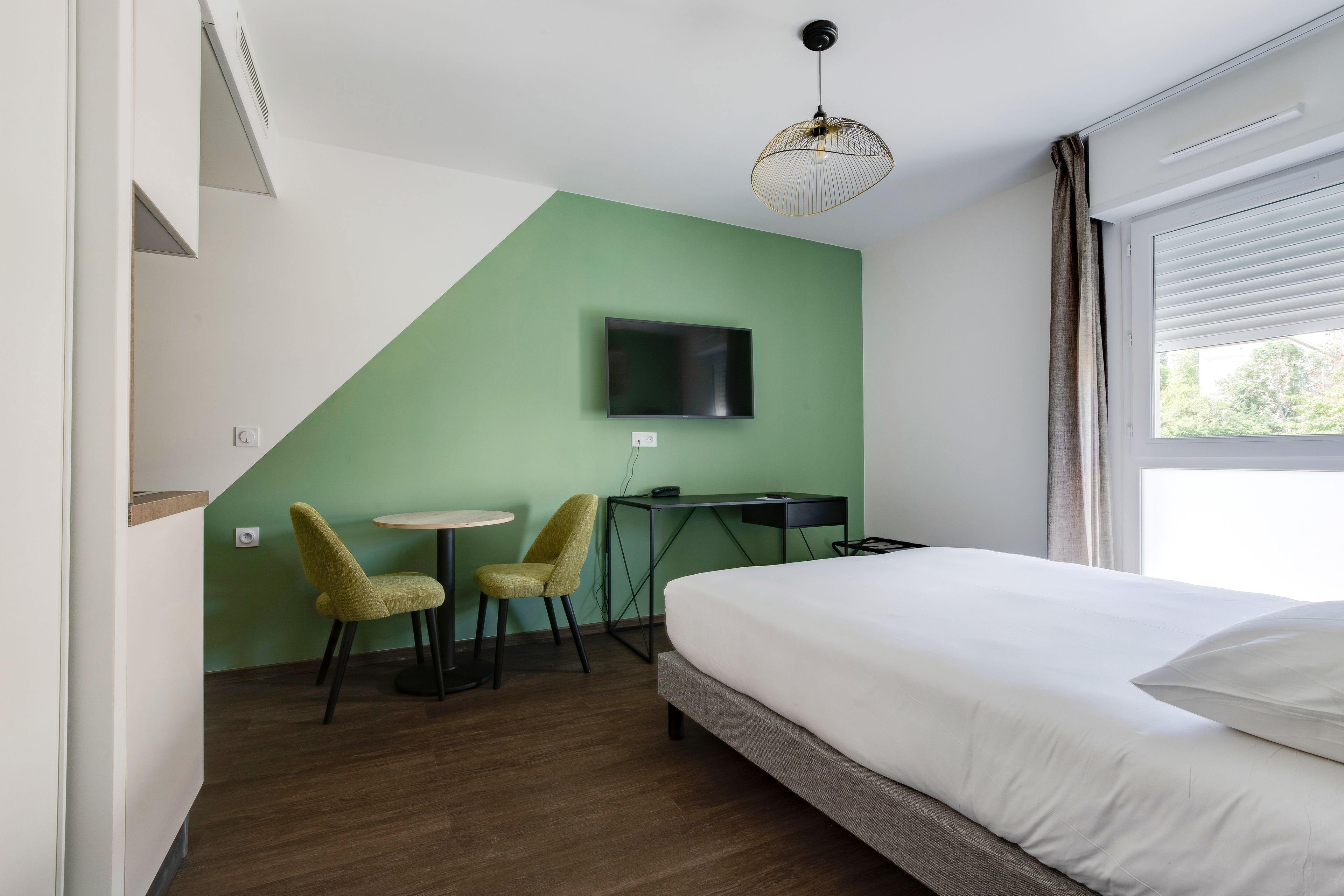 A hotel room with a bed, a desk, two chairs, a TV mounted on the wall, and a window with curtains.