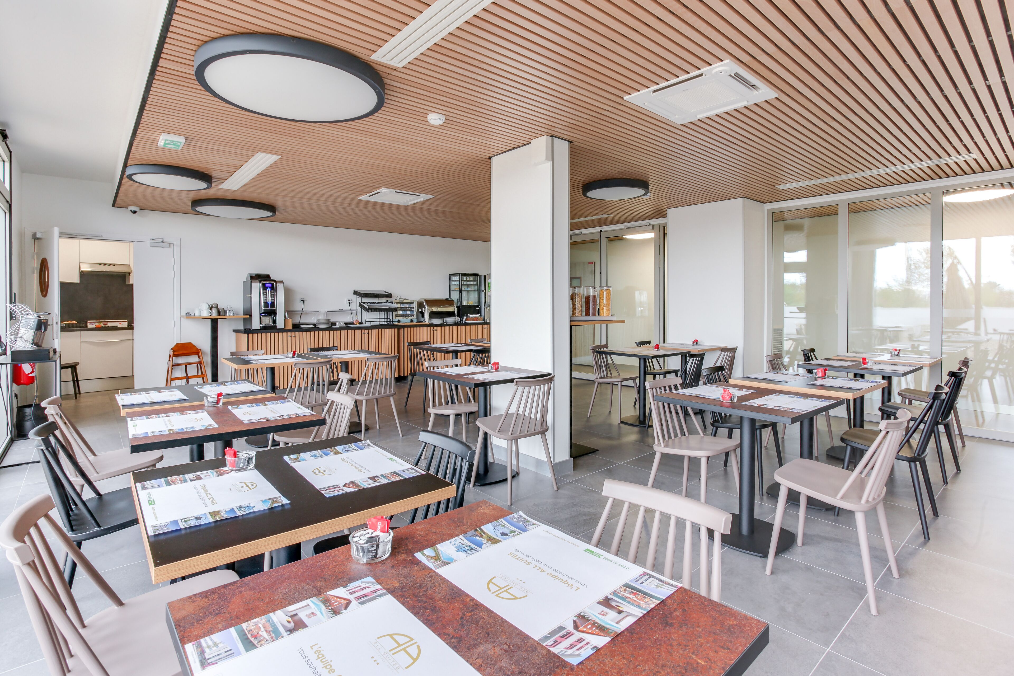 Um restaurante moderno com painéis de teto em madeira, várias mesas com cardápios, e uma área de cozinha visível ao fundo.