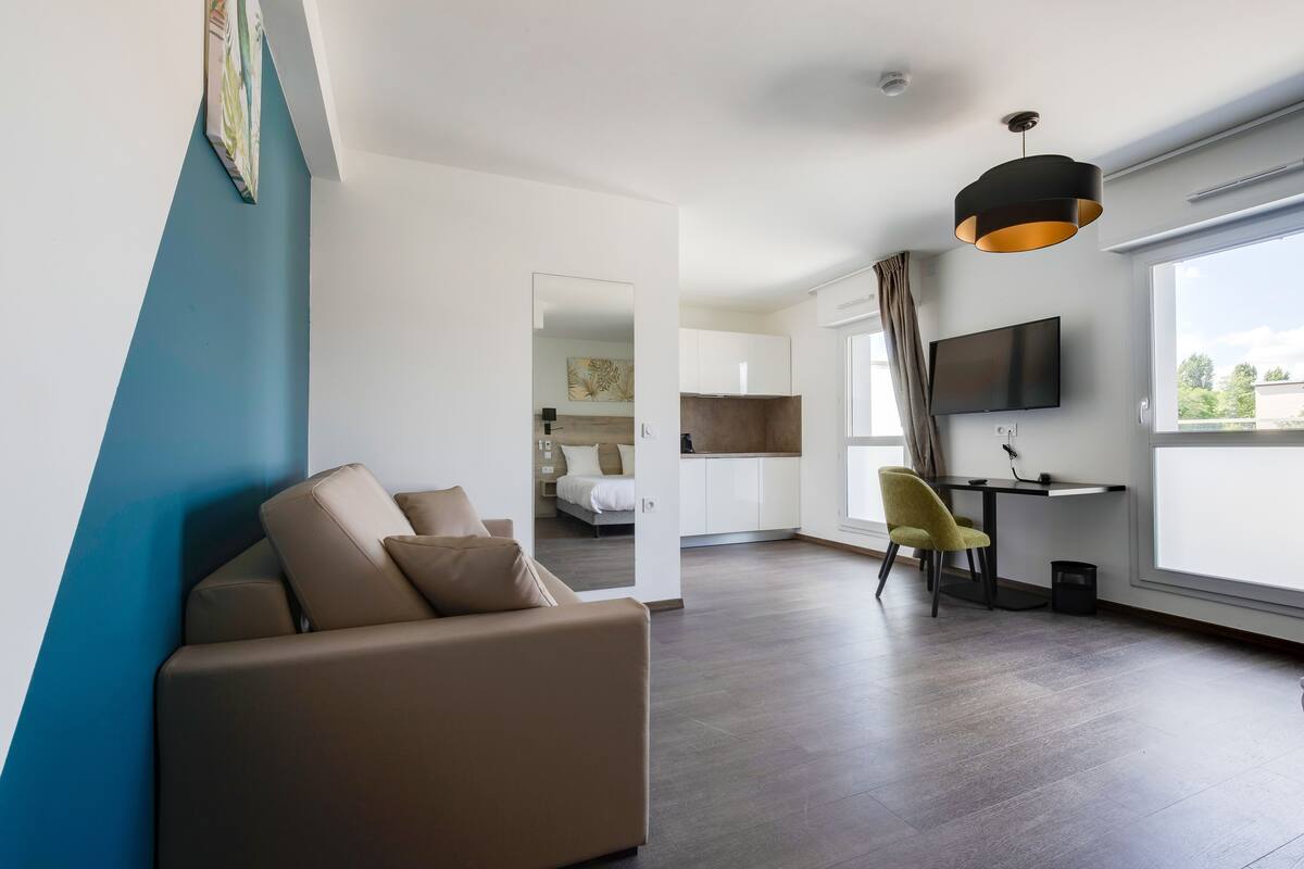 A modern living room with a sofa, a desk, and a TV. There is a window with curtains and a view of the outside.