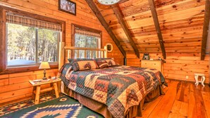 A cozy wooden cabin bedroom with a large bed, a nightstand, a dresser, and a window with a view of trees. - Webb Cabin Mountain (Angel Fire)
