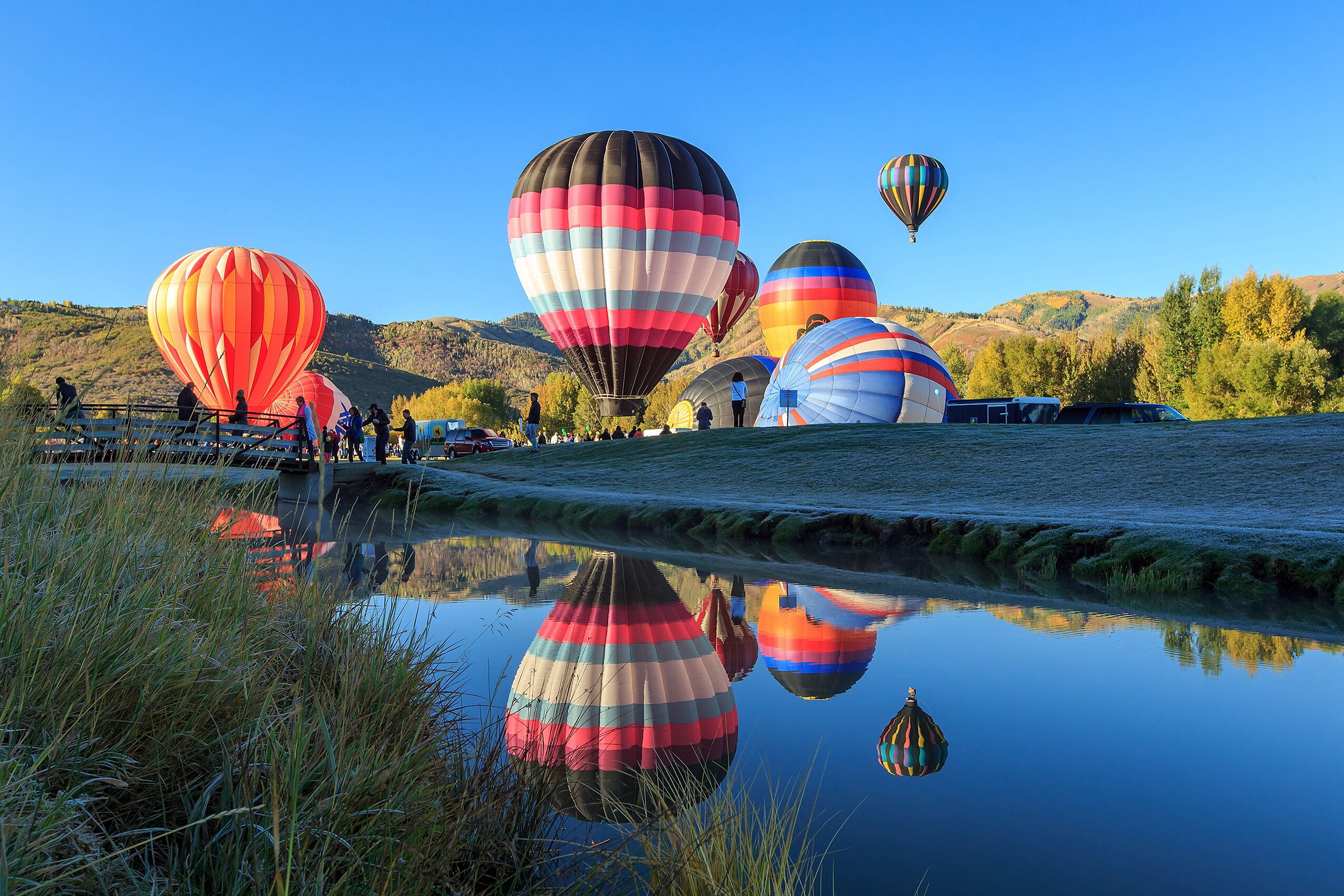 A group of hot air balloons over a calm body of water with reflections, surrounded by grassy areas and hills.