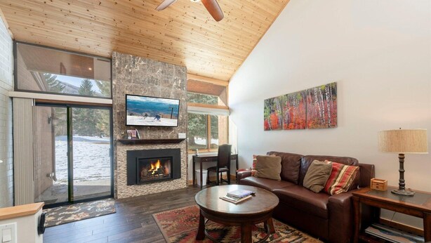A modern living room with a stone fireplace, a leather sofa, and a large window overlooking a body of water. - Stylish Park City 2BR Sleeps 6-Walk to Park City Resort, Shops & Dining (Park City)