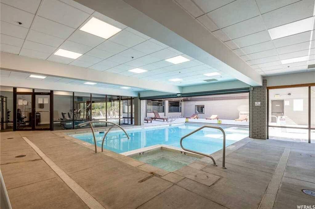 An indoor swimming pool with clear glass walls, lounge chairs, and a tiled floor.