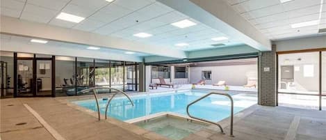 An indoor swimming pool with clear glass walls, lounge chairs, and a tiled floor.