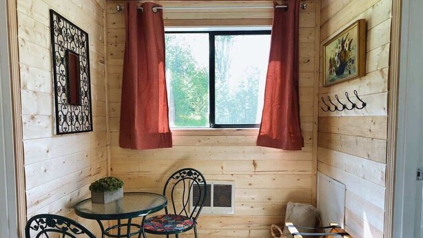 Une petite pièce chaleureuse aux murs en bois, avec une table ronde entourée de chaises, une fenêtre avec des rideaux, un radiateur et une petite plante.
