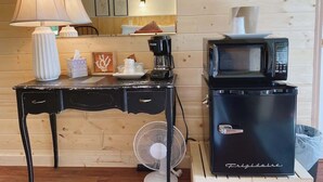 Une petite cuisine équipée comprenant un réfrigérateur noir, un four à micro-ondes, une machine à café, un ventilateur et une petite table agrémentée d’une lampe et d’objets décoratifs.
