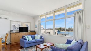 A living room with a sofa, a coffee table, and a TV. There is a window with a view of a body of water and buildings. - Mandurah Overlooking The Marina (Mandurah)