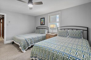 Une chambre avec deux lits, un ventilateur de plafond, une fenêtre avec des stores et un tableau encadré au mur.