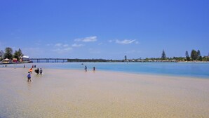 Ein Strand mit klarem Wasser, ein Steg im Hintergrund und Menschen, die am Ufer entlanggehen.