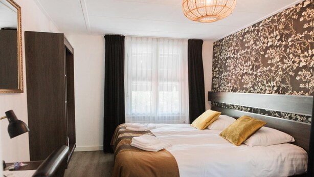 Een hotelkamer met een bed, een bureau, een stoel, een lamp en een groot raam met gordijnen.
