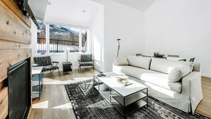 A modern living room with a white sofa, a coffee table, and a patterned rug. There is a TV on the left and a window with a view of mountains. - Stylish Park City 2BR, Walk to Three Kings Lift, Heated Pool & Hot Tub (Park City)
