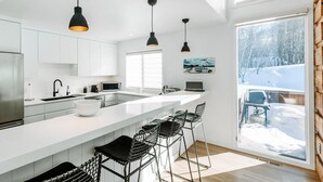 A modern kitchen with white cabinets, a large island with bar stools, pendant lighting, and a view of a snowy landscape through a large window. - Stylish Park City 2BR, Walk to Three Kings Lift, Heated Pool & Hot Tub (Park City)