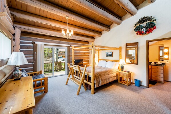 Quarto de cabana de tronco com cama de madeira, escrivaninha, cadeira, mesa pequena, mesa de cabeceira e banheiro com espelho e pia.