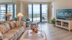 Hotelkamer met een bank, een bijzettafeltje met bloemen, een televisie en een balkon met uitzicht op zee.