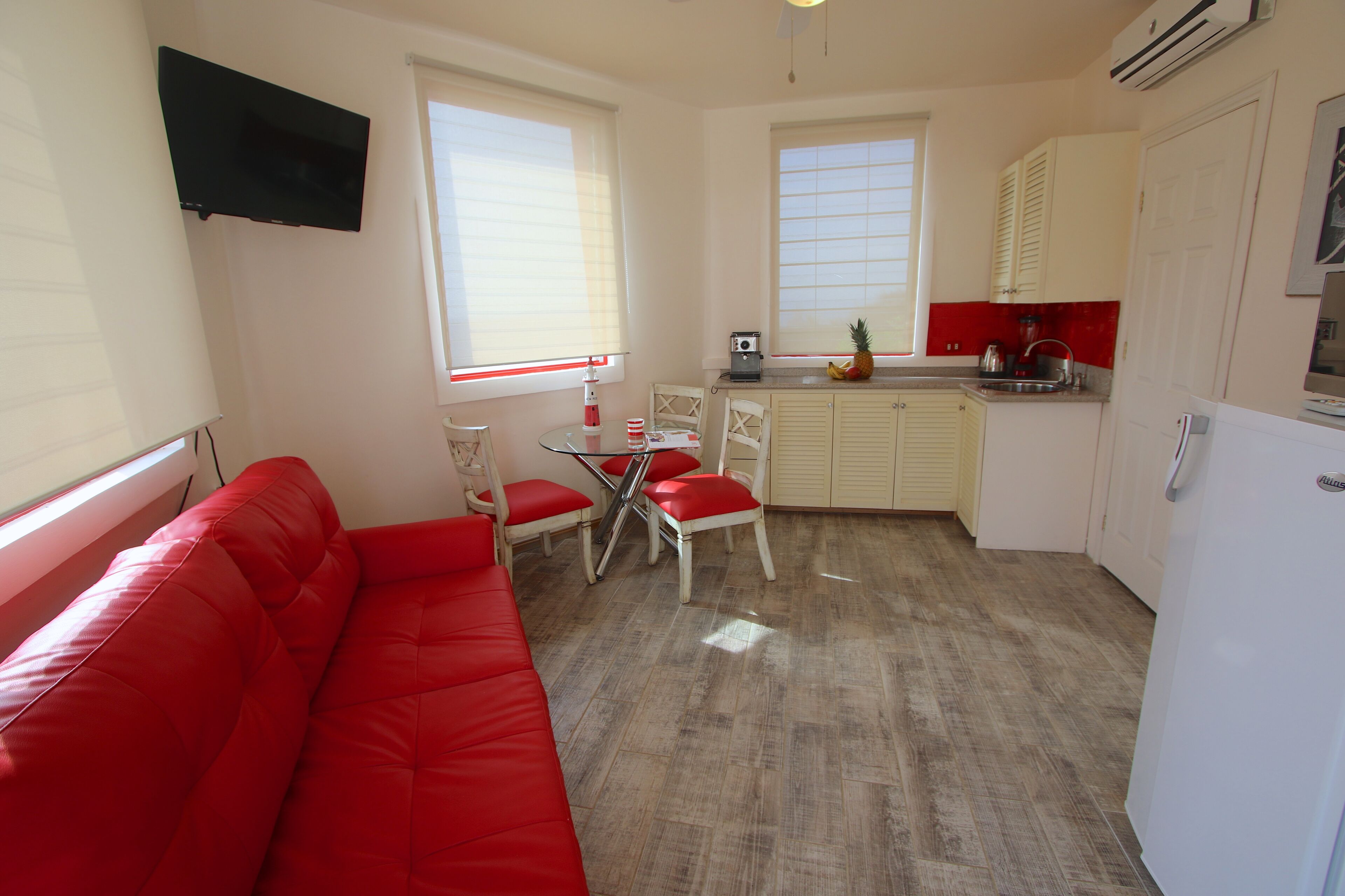 A compact living space with a red sofa, a dining table with chairs, a kitchen area with cabinets, and a flat-screen TV mounted on the wall.