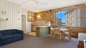 A compact kitchen with a refrigerator, microwave, and sink. There is a dining area with a table and chairs, and a blue sofa. A window provides a view of the outside. - The Sands, Unit 5 - The Entrance, NSW (The Entrance)