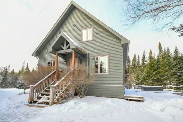 A two-story house with a wooden staircase leading to a front porch, surrounded by snow and trees.